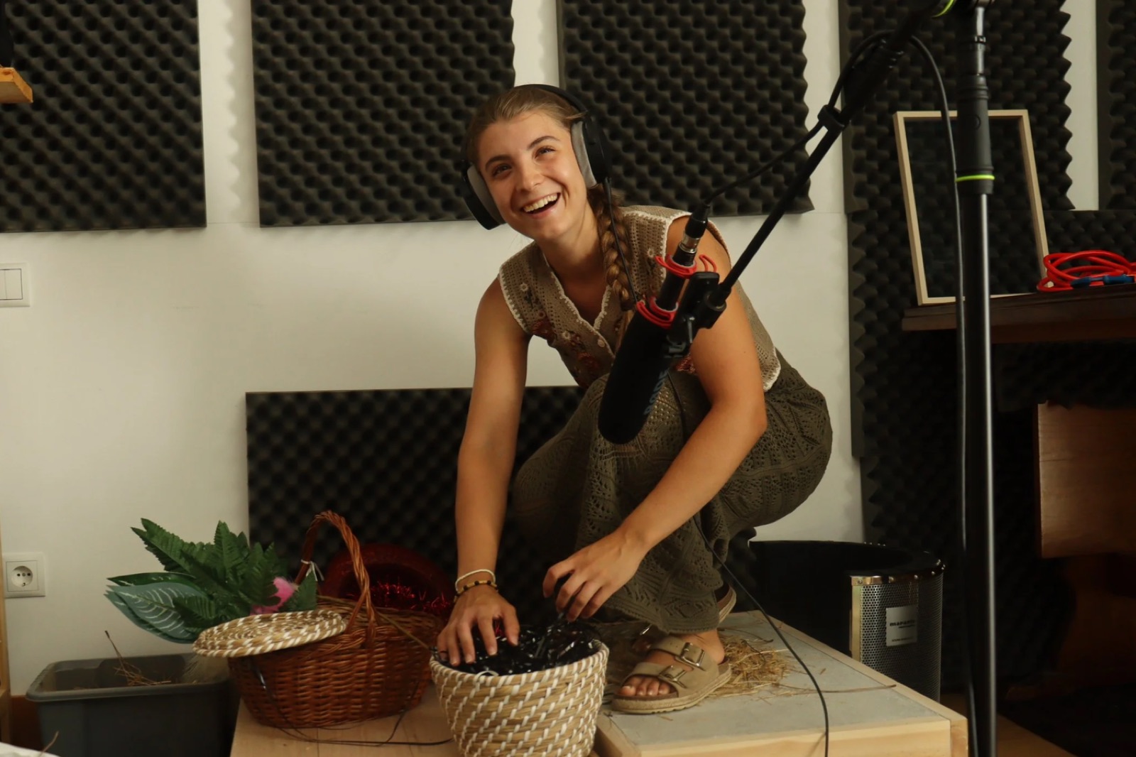 Lúcia Carvalho wearing headphones, crouched over a woven basket of props in front of a boom microphone, laughing.